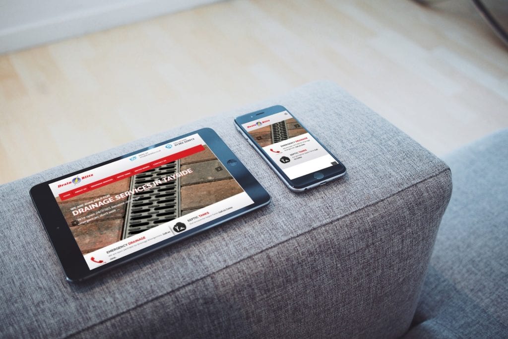 A tablet and phone sitting on the arm rest of a modern grey sofa displaying the DrainBlitz website